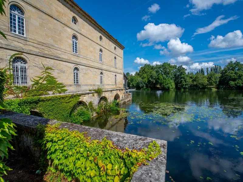 Porchères Mill in Bordeaux, a stone building with regular facades overlooking a calm river, surrounded by lush green vegetation and poplar trees under a blue sky.