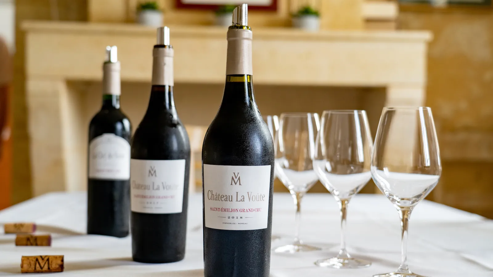 Three bottles of red wine Château La Voûte Saint-Émilion Grand Cru vintage 2018 lined up on a white table with empty glasses and cork stoppers bearing the M logo, in an elegant Bordeaux interior.