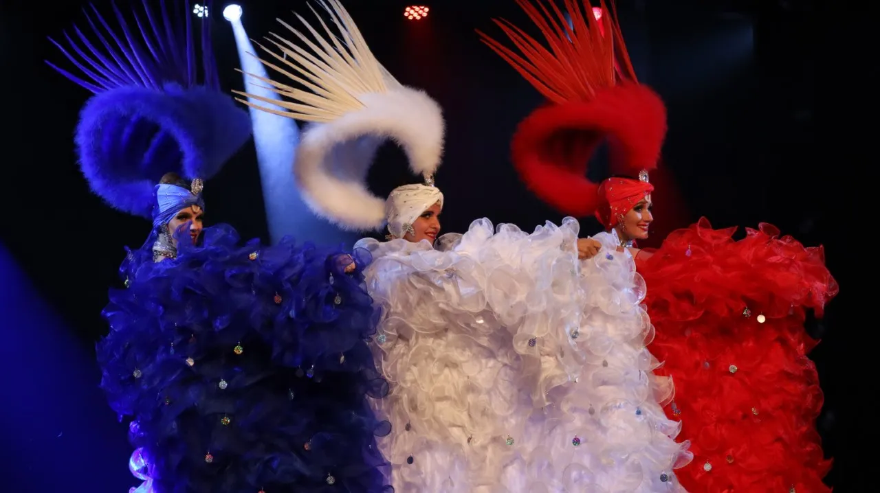Three cabaret dancers on stage wearing elaborate blue, white, and red costumes with ornate headdresses and voluminous feathered backpieces, lit by colored spotlights at Le Grain d'Folie in Bordeaux.