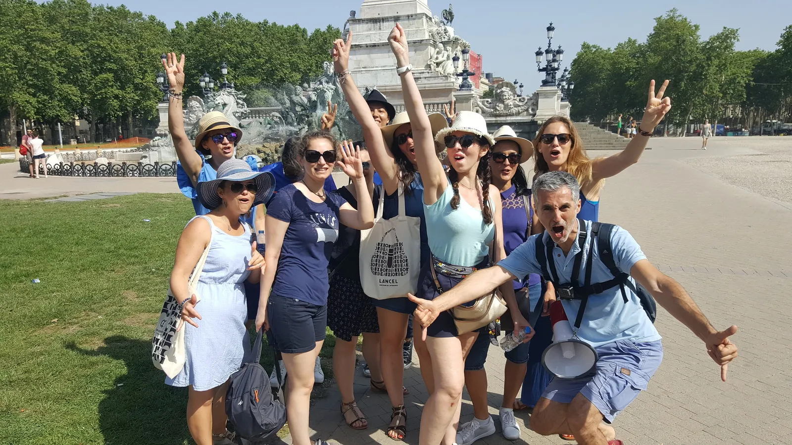 A cheerful group of tourists posing in front of the Monument of Place de la Bourse in Bordeaux, raising their arms in celebration during a guided tour.