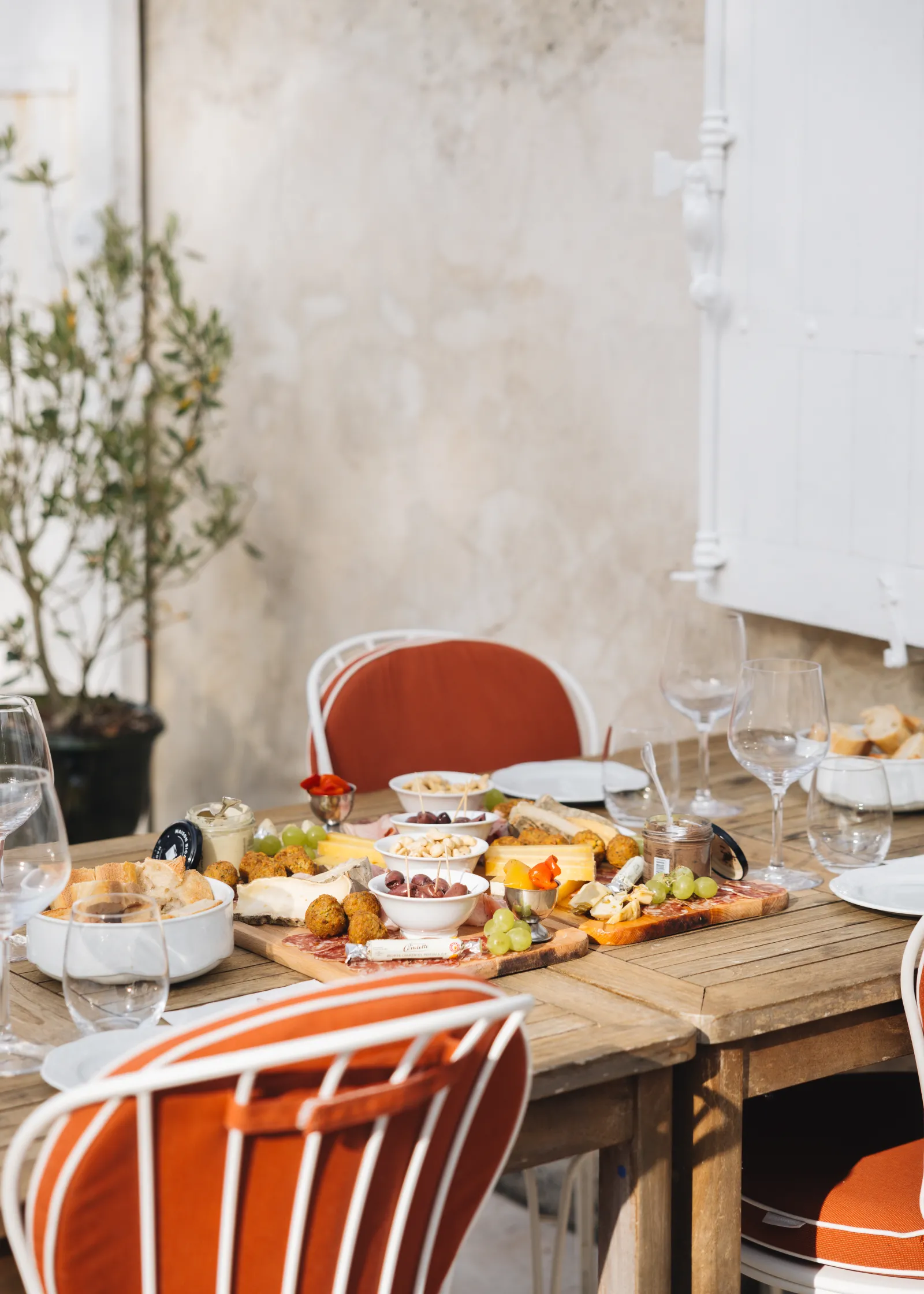 Dining table set with orange and red chairs, charcuterie and cheese board, glasses and plates, potted plant in background, light grey walls