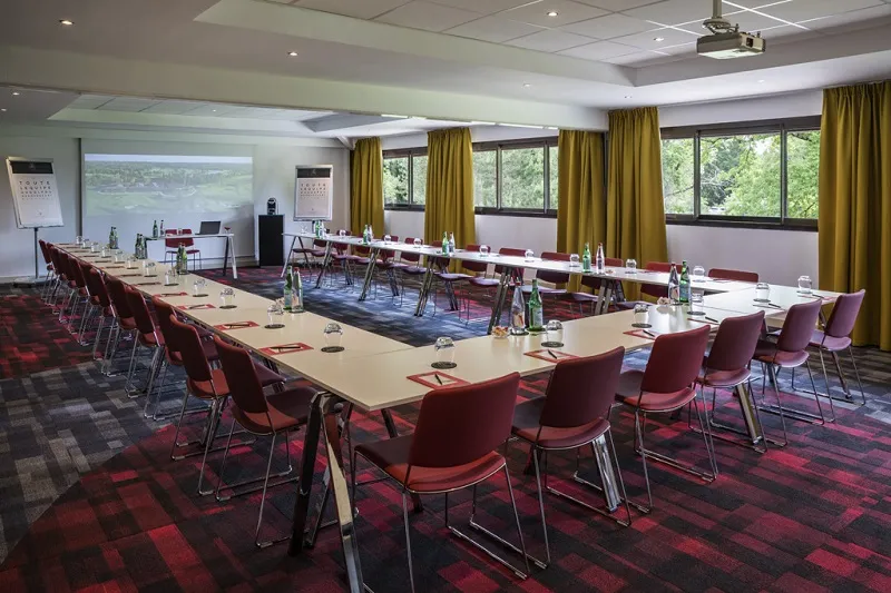 Modern conference room at Cabot Bordeaux MGallery Collection hotel featuring long rectangular tables, burgundy red chairs, golden curtains, projection screen, and large windows overlooking green surroundings.