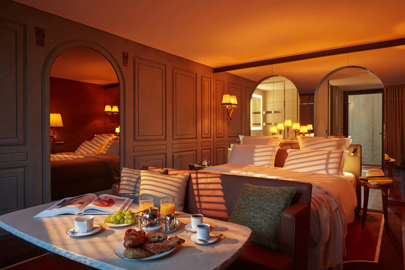 Luxury suite at Mondrian Bordeaux Les Carmes featuring a king-size bed, arched mirrors, warm golden lighting, and breakfast service on a low table