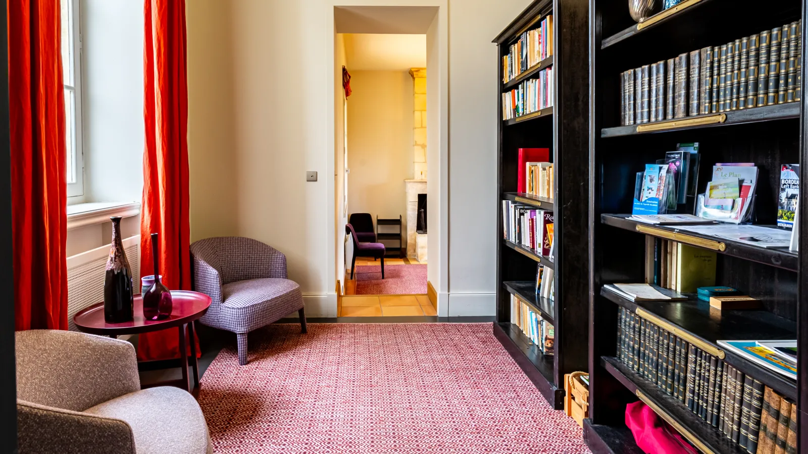 Elegant living room at Château Malescasse in Bordeaux featuring a black bookshelf, gray armchairs, red curtains, and access to an adjacent room with yellow walls.