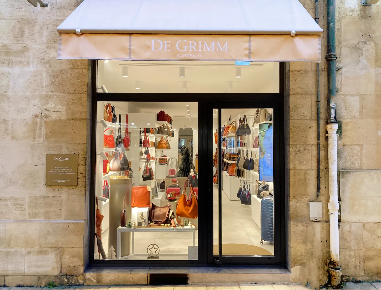 De Grimm luxury handbag and accessories store in Bordeaux, featuring an illuminated storefront displaying leather goods in warm tones.