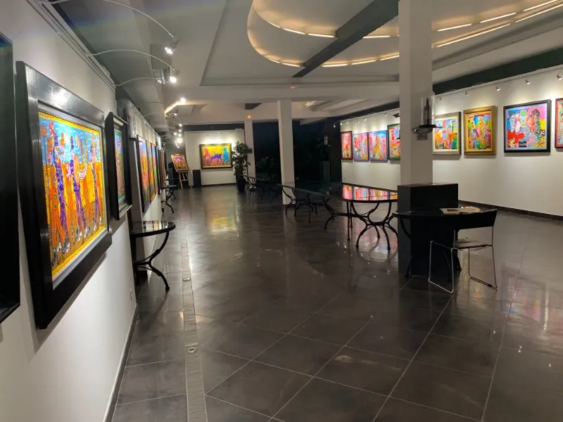 Modern art gallery featuring colorful paintings displayed on white walls, polished gray flooring, black furniture, contemporary ceiling with integrated lighting.