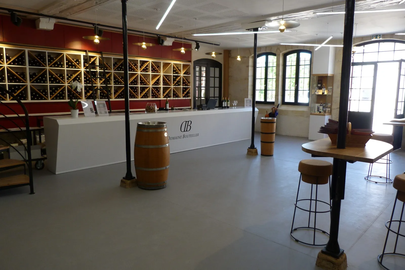Modern interior of a wine tasting room with Domaine Boétellers counter, golden wine racks, wooden barrels, high tables and large windows in Bordeaux.