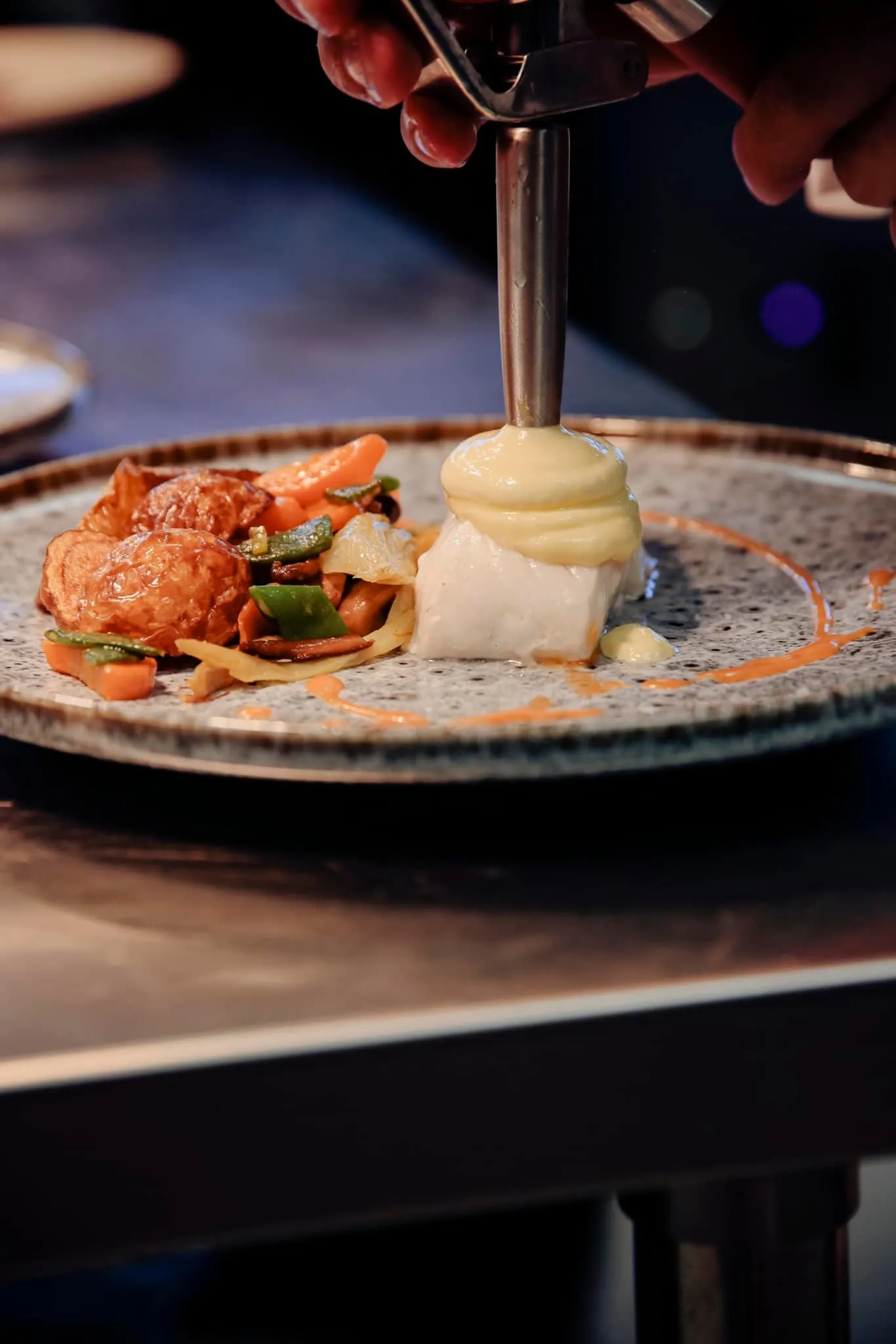 A chef pours sauce over a gourmet dish presented on a gray plate, featuring lobster, vegetables, and white mousse, at Magnus restaurant in Bordeaux.