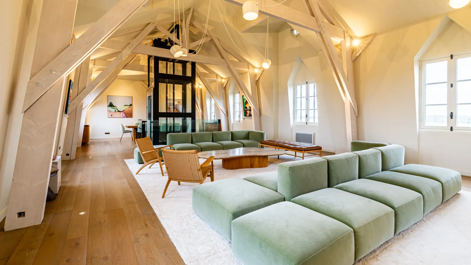 Bright spacious interior with cathedral-style white beam ceiling, modern sage green and rattan furnishings, wooden coffee tables, large windows, and central black staircase at Château Malescasse in Bordeaux.