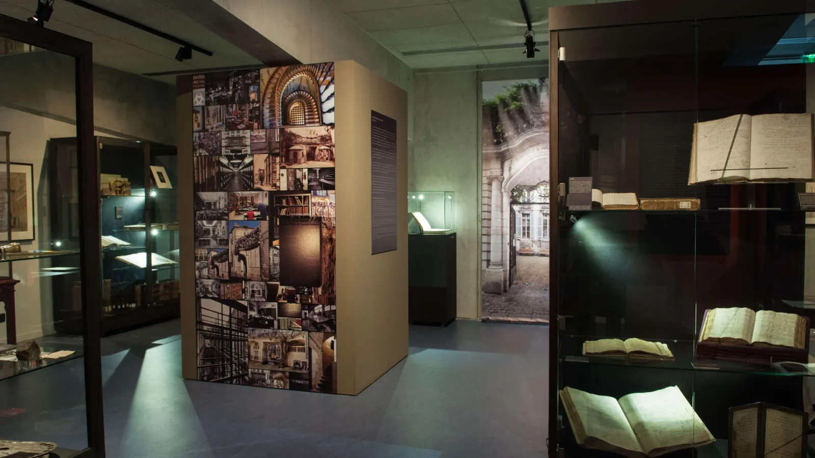 Interior of a museum exhibition showcasing Bordeaux's architectural heritage, featuring display cases with historical artifacts, photographic panels of urban architecture, and antique books illuminated with directional lighting.