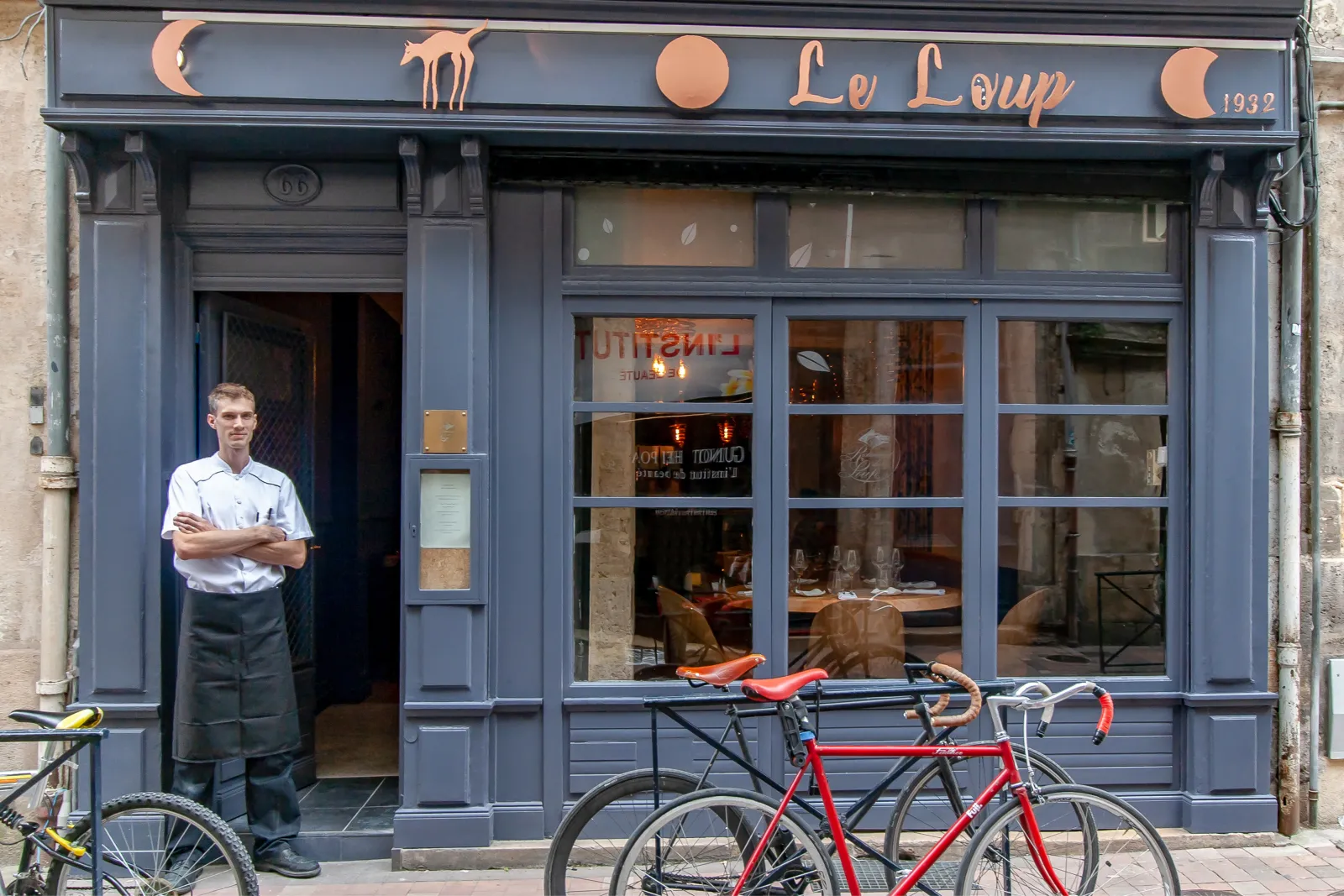 Storefront of Le Loup restaurant in Bordeaux with a waiter in professional attire at the entrance and red bicycles parked outside