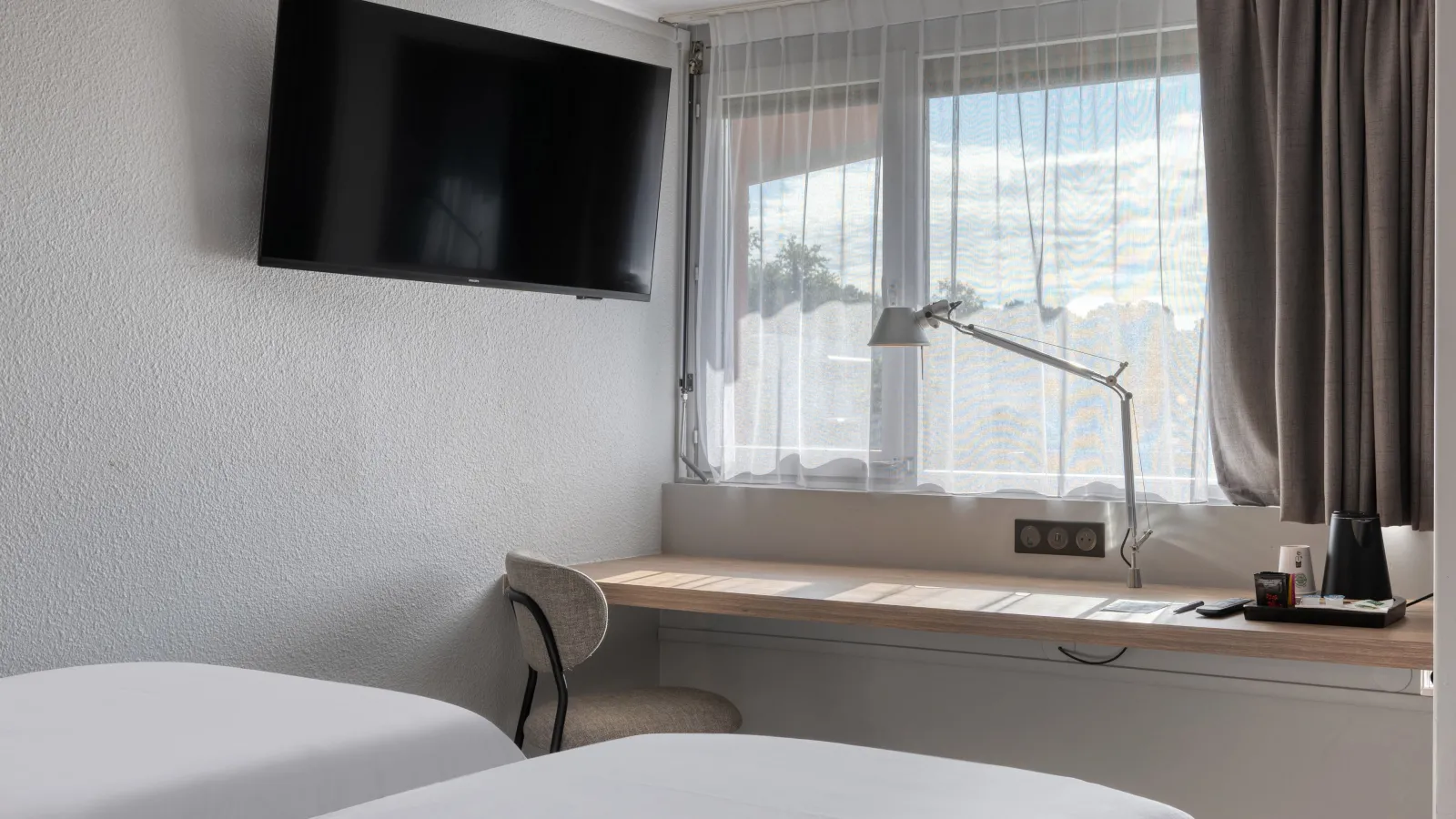 Modern hotel room featuring a white bed, wall-mounted TV, and desk area overlooking panoramic views of Bordeaux through large windows. Black desk lamp and beige chair complete the setup.