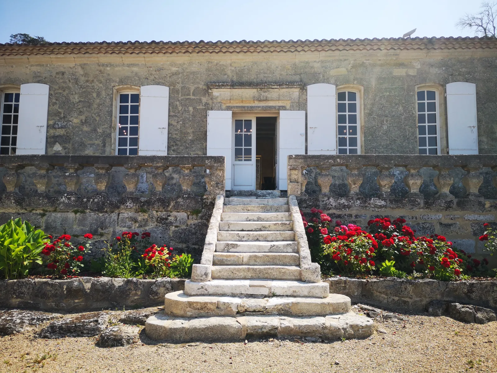 Historic stone façade of an old building with white doors and shutters, central staircase and red flowers bordering the entrance, located in Bordeaux.