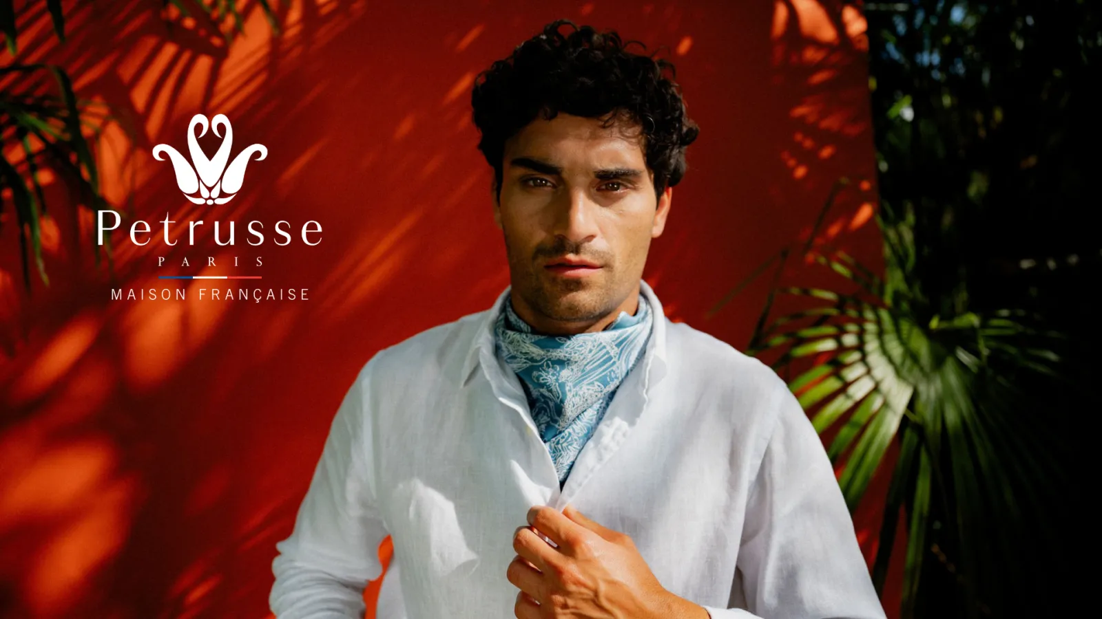 Man wearing white shirt and blue patterned scarf against red background with tropical foliage, Petrusse Paris French House logo