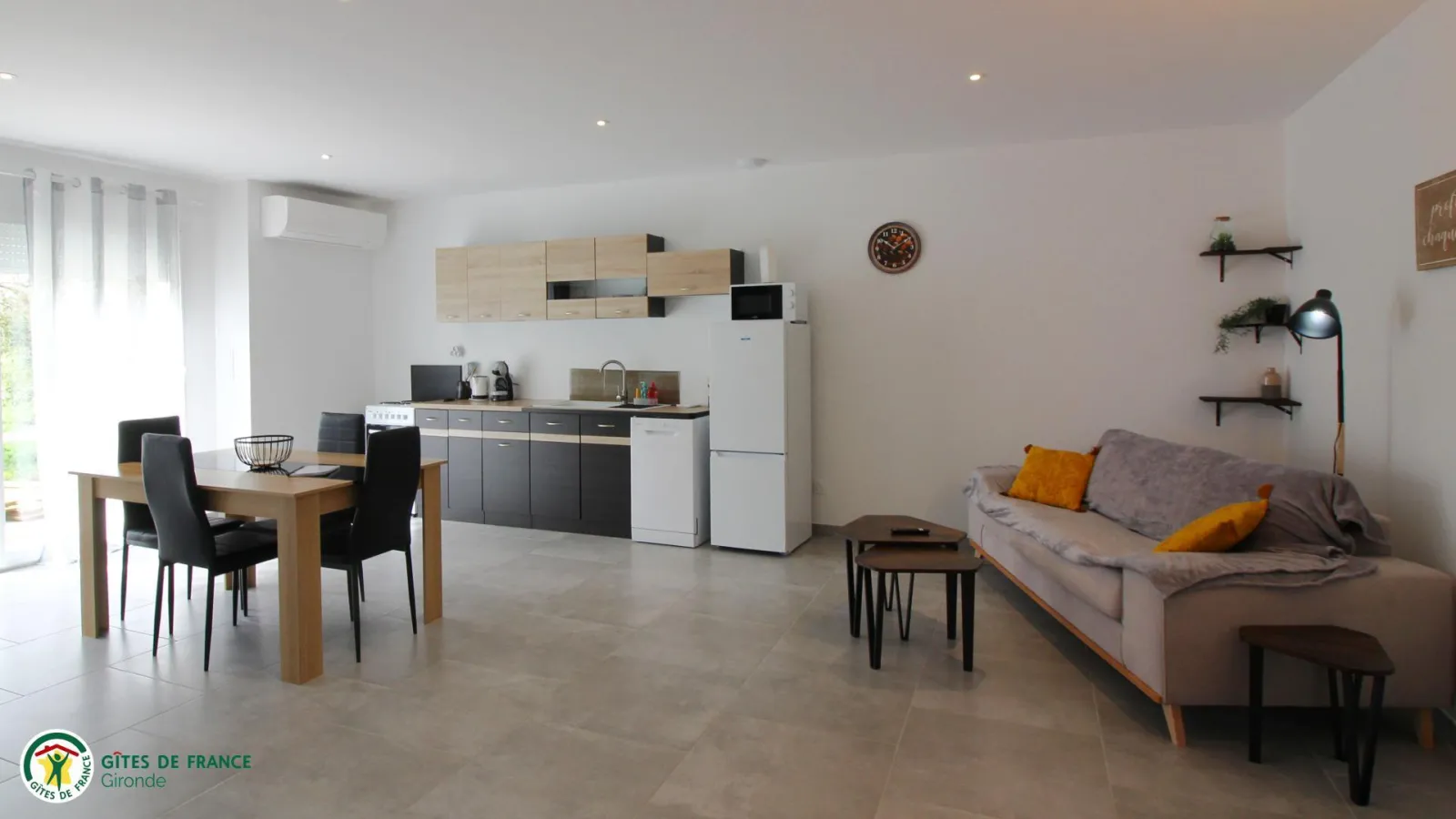 Modern interior of a holiday rental featuring equipped kitchen, wooden dining table, grey sofa with yellow cushions and contemporary decor, located at Château Pierre de Montignac in Bordeaux.
