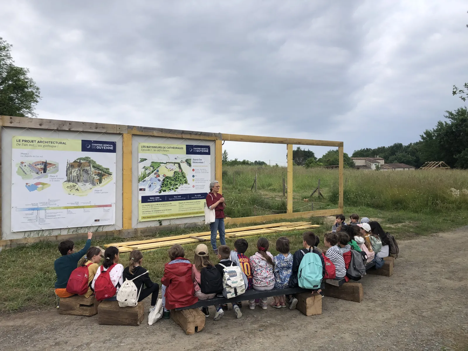 A group of children sitting on wooden benches listen to a presenter explaining information panels about the Guyenne medieval site in Bordeaux, with architectural plans and garden illustrations visible on the display boards.