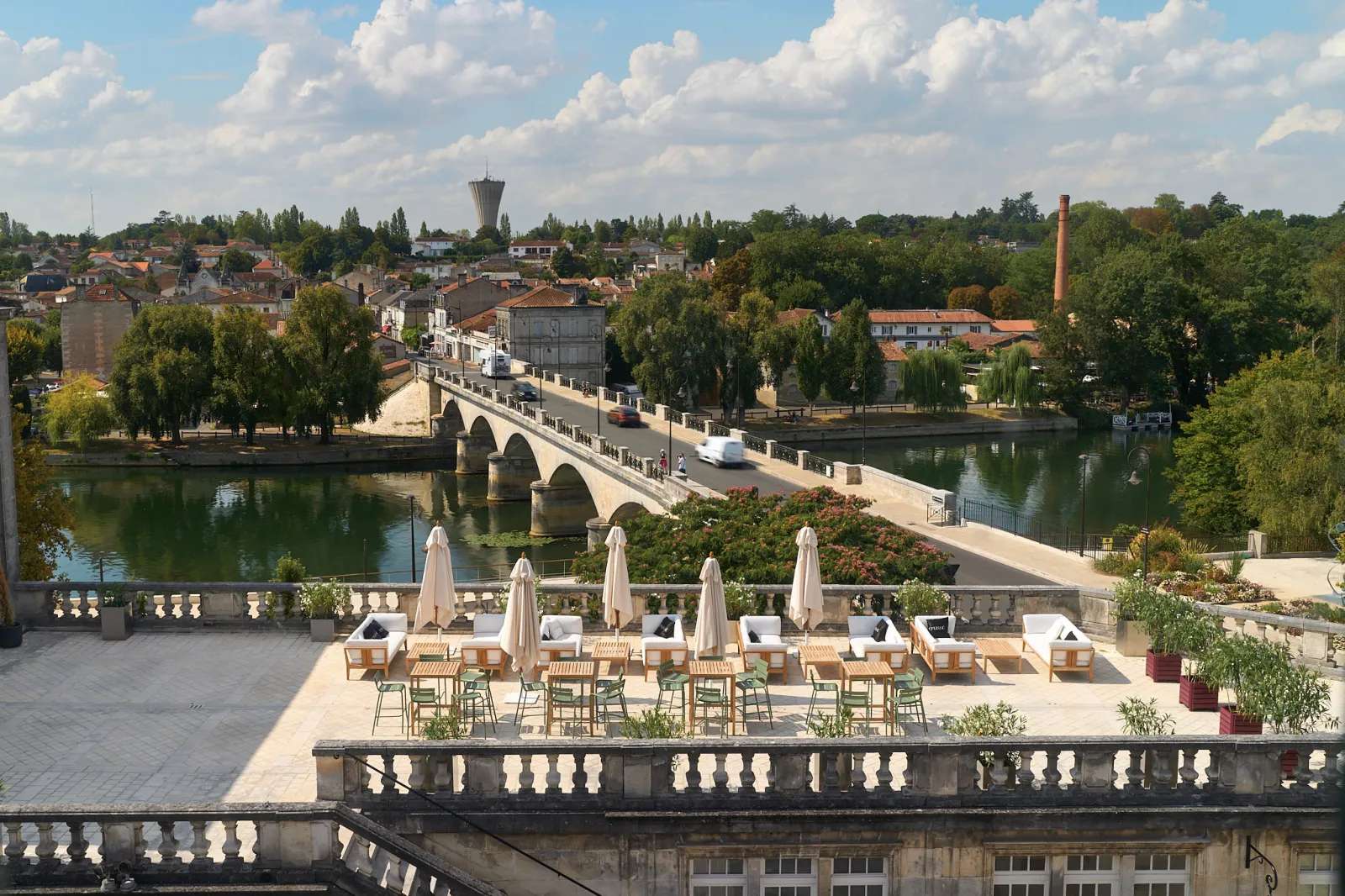 Terrace with sun loungers and umbrellas overlooking a stone bridge spanning a river, with a historic town and old buildings in the background