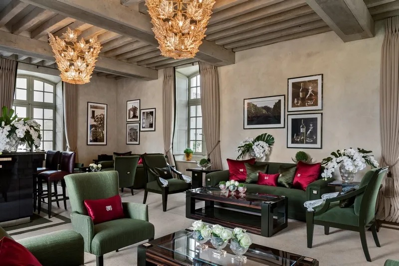 Elegant lounge at Lalique Hotel in Bordeaux featuring green armchairs, red cushions, ornate gilt crystal chandeliers, exposed ceiling beams, and framed artworks
