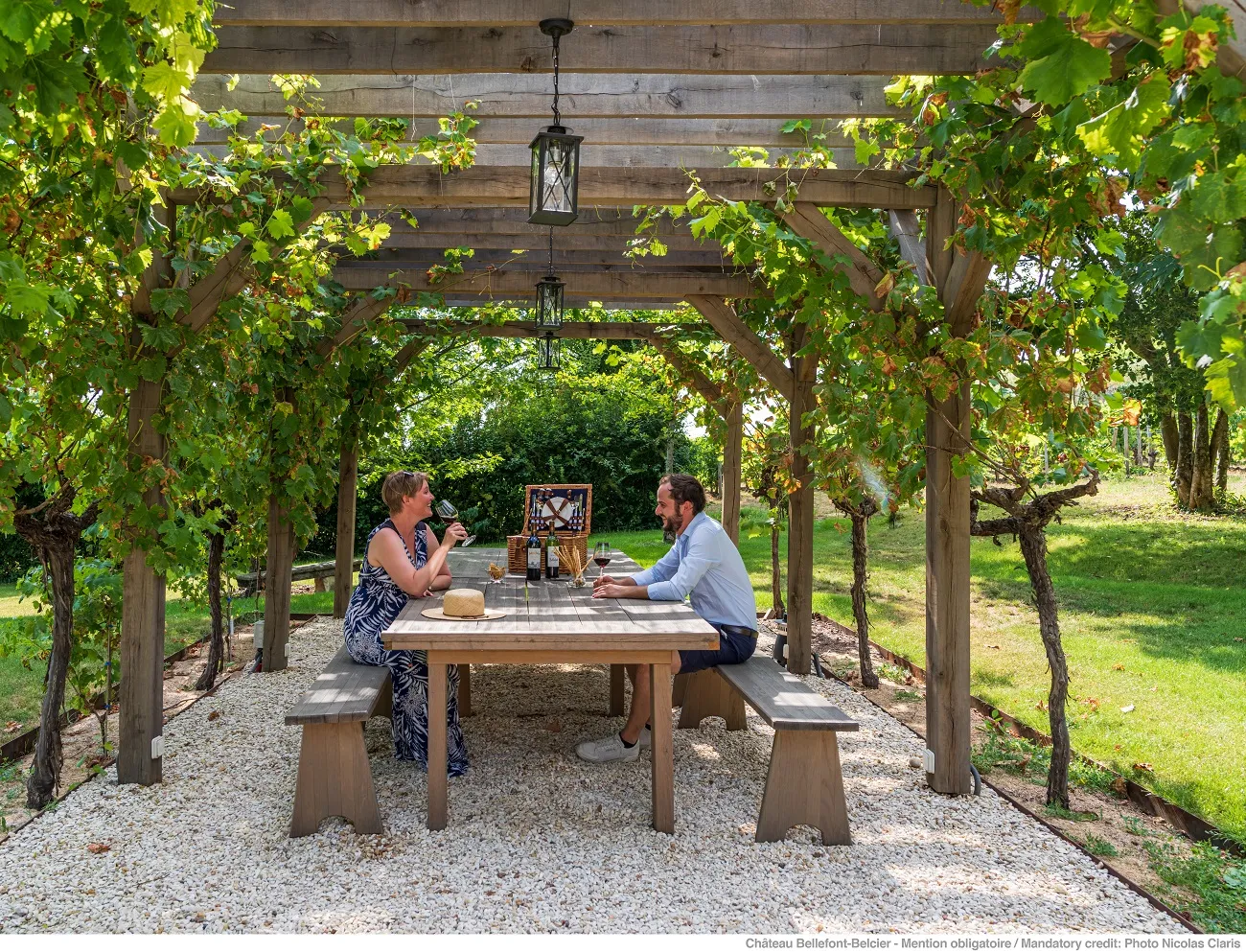Two people enjoy wine tasting at a wooden table under a vine-covered pergola at Château Bellefont-Belcier in Bordeaux, with glasses and a bottle on the table.