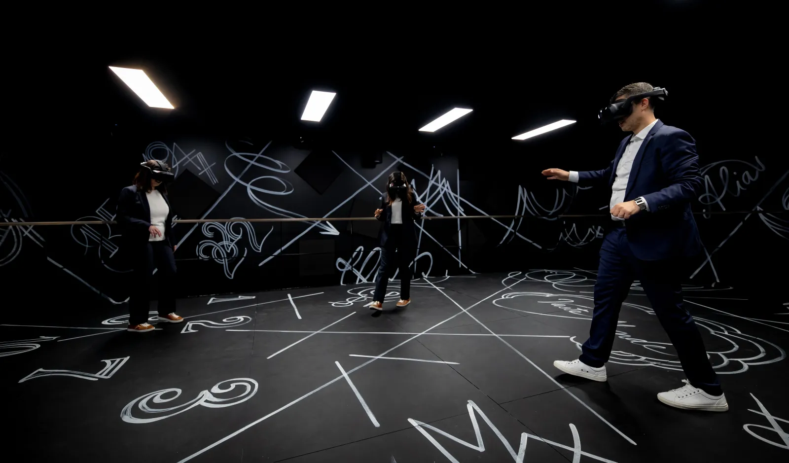 Immersive tour at Hennessy House in Bordeaux: a man in a blue suit and three visitors wearing virtual reality headsets explore an interactive black room decorated with luminescent white patterns on the floor and walls.
