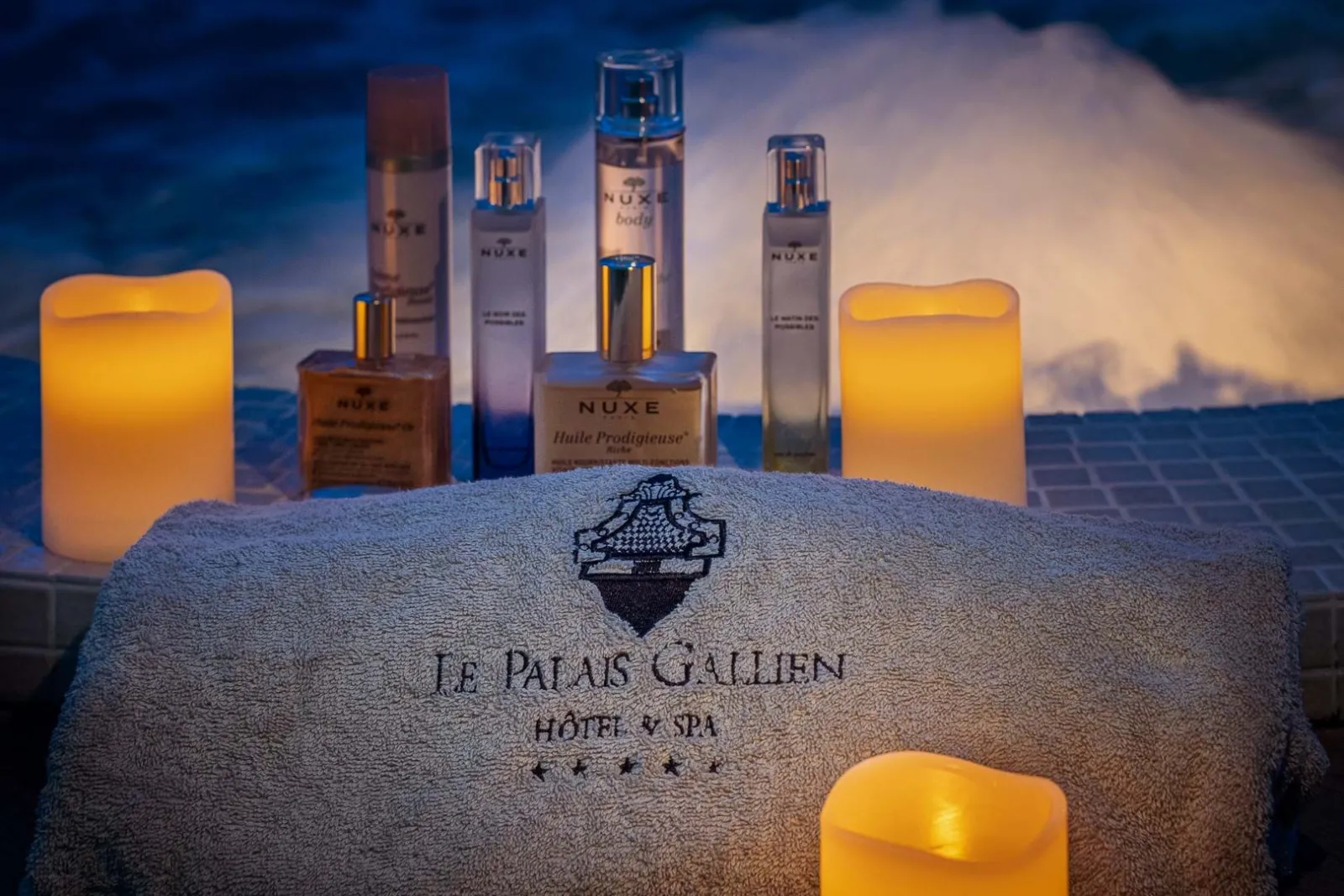 Palais Gallien Hotel & Spa towel displayed with NUXE beauty products and lit candles against a sunset backdrop in Bordeaux.