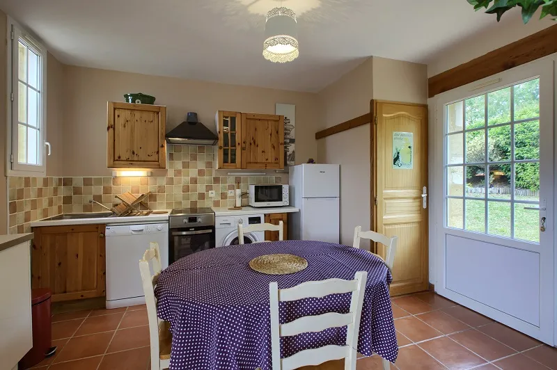 Bright spacious kitchen at Château La Gontrie featuring a round table with purple polka dot cloth, wooden cabinets, beige checkered tile, white appliances, and French doors opening to a lush green garden.