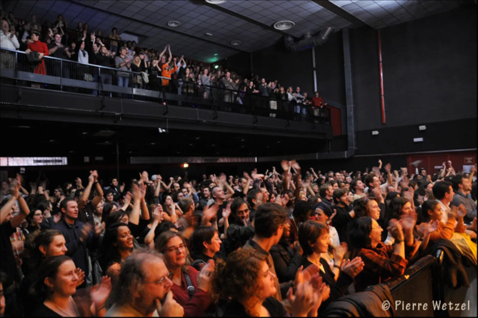 Enthusiastic crowd at a concert at Krakatoa in Mérignac, Bordeaux, with spectators applauding in the venue and fans in the upper seating area.