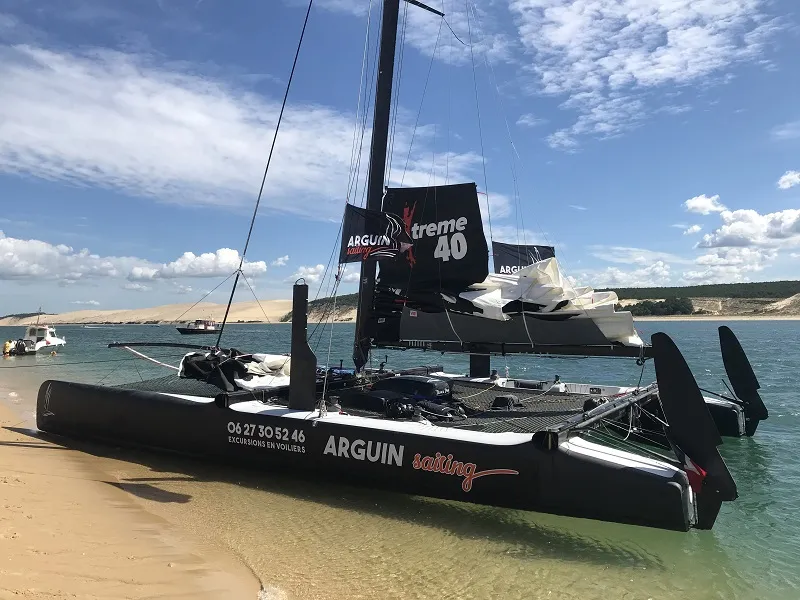 Black Arguin Xtreme 40 catamaran beached on sand with black sails, moored near a rocky coastline