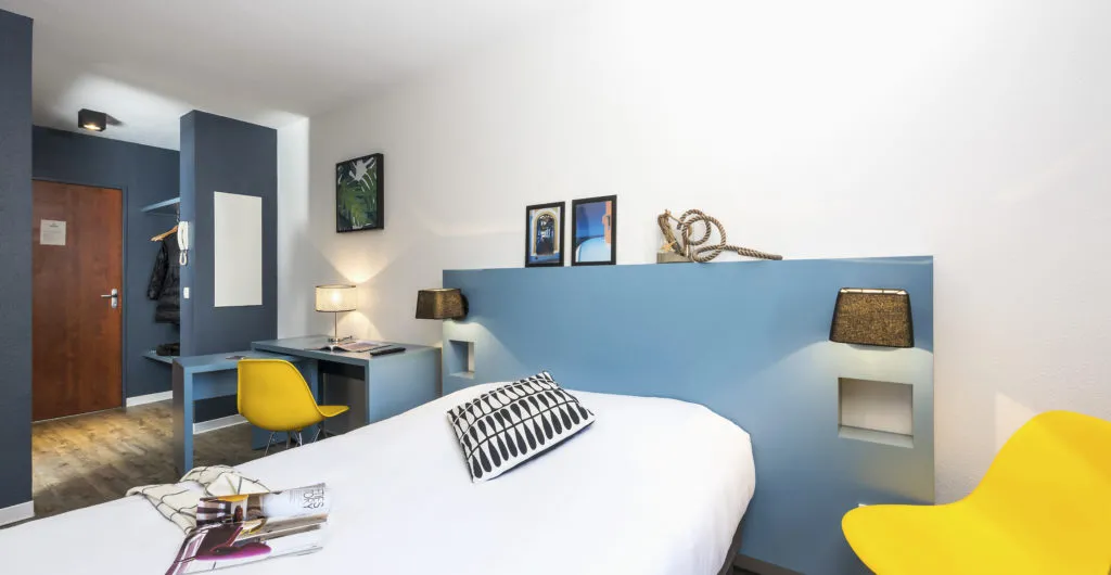 Modern bedroom at Ténéo Apparthotel Bordeaux Saint-Jean featuring a white bed, blue headboard, yellow chairs, integrated desk, and minimalist décor.
