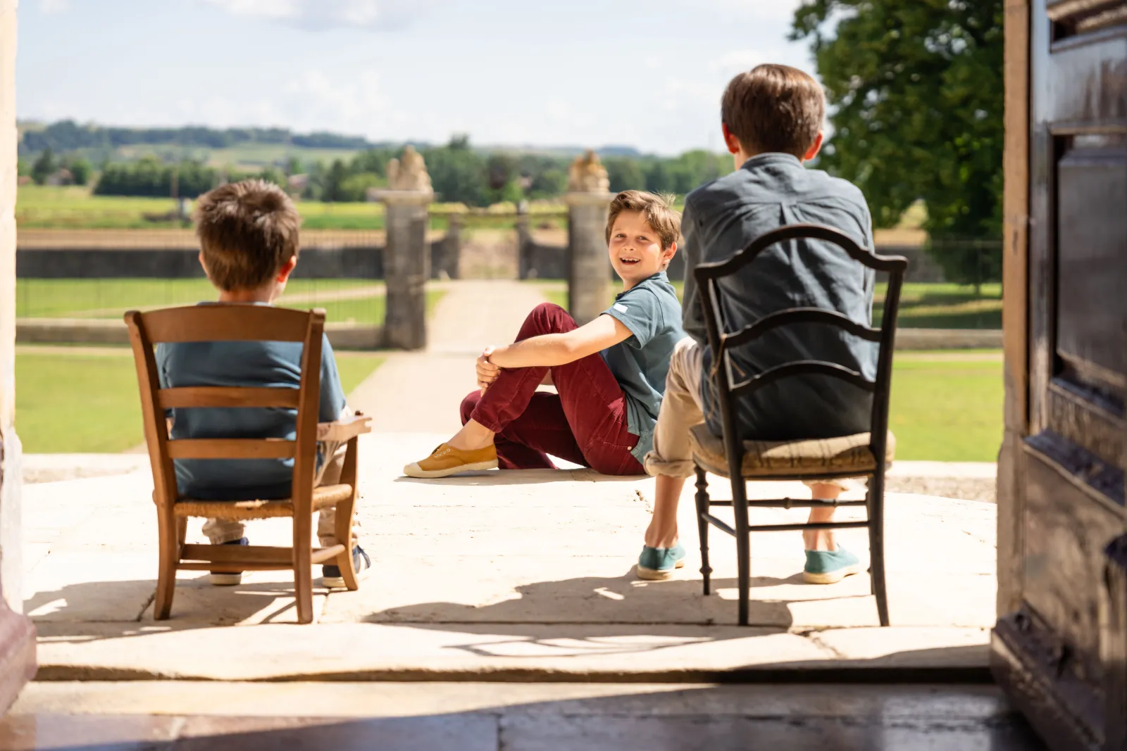 Three children sitting on chairs overlooking the Bordeaux countryside, with the Château de Malle gardens and architecture visible in the background on a clear day.