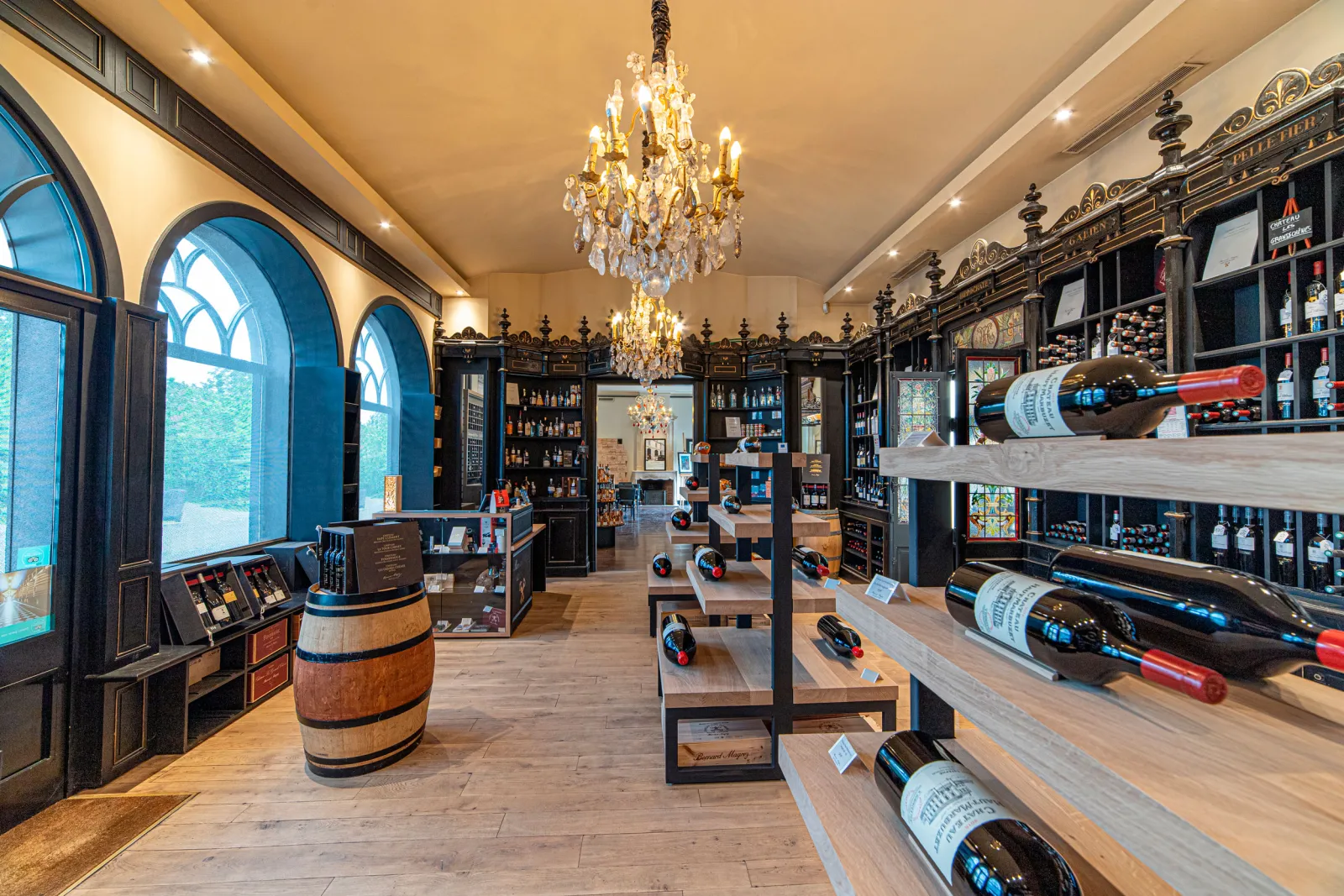 Elegant interior of a wine cellar displaying Bordeaux bottles in wooden displays, crystal chandelier, oak barrels, and blue arched windows at Château Pape Clément.