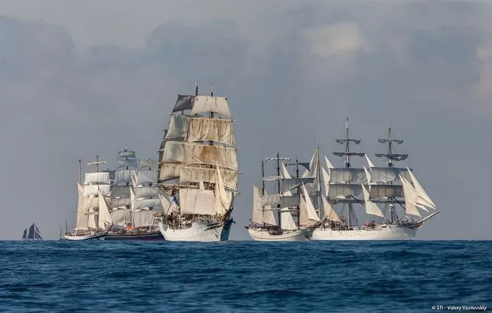 Three traditional sailing ships with white sails sailing together on blue ocean waters during the Tall Ships Races in Bordeaux.
