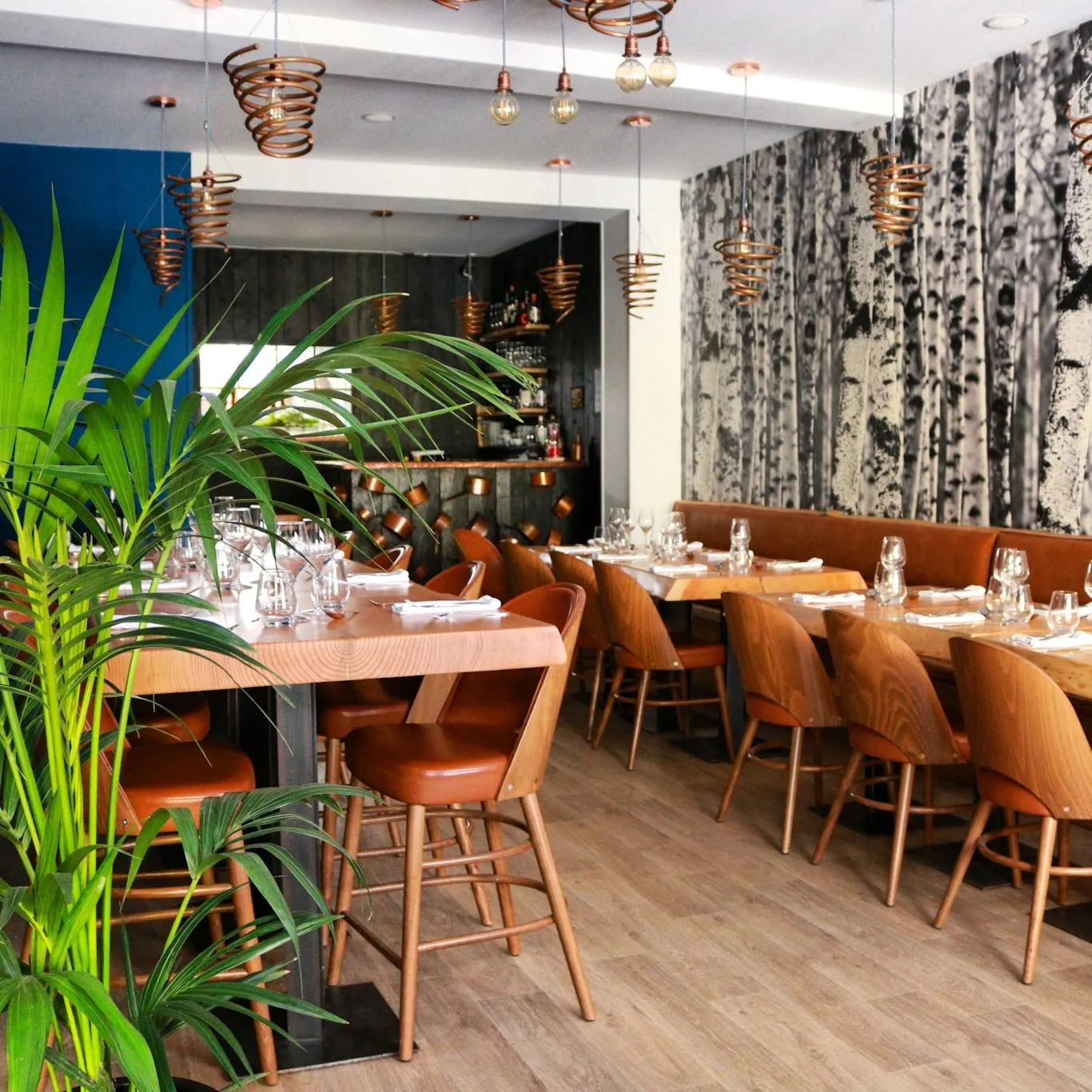 Modern interior of Le Loup restaurant in Bordeaux featuring wooden tables, cognac leather chairs, suspended gold fixtures, blue and grey walls, green plants and bar in the background