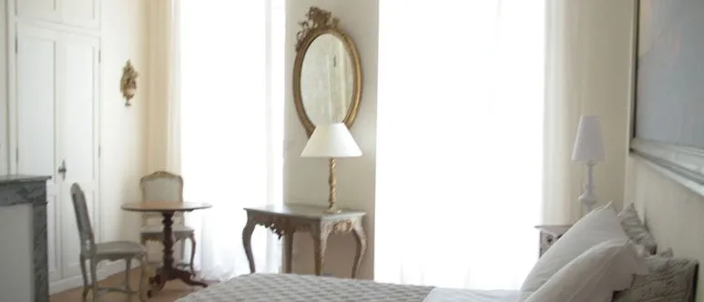 Elegant classical-style bedroom featuring a white bed, ornate mirror above a console table, wooden chair, table lamp and large windows with natural light