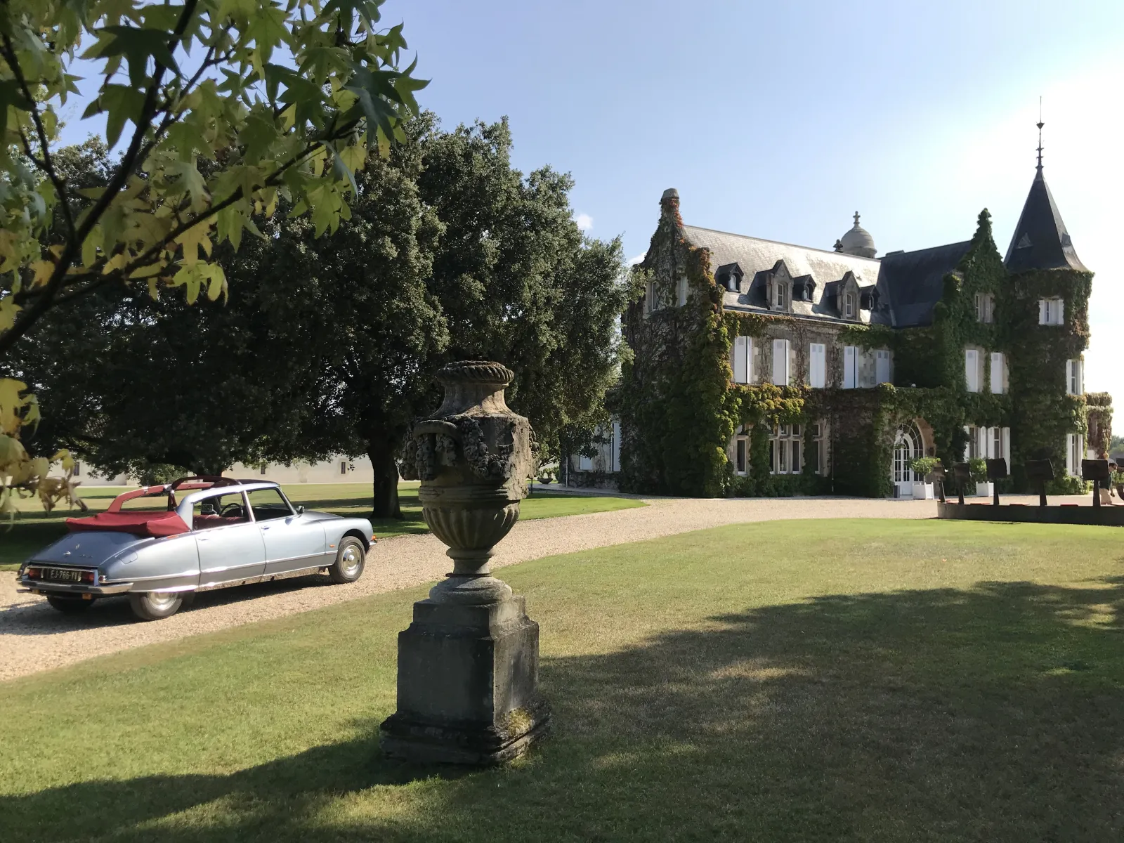 A classic white and red convertible car parked in front of a 19th-century stone château with turrets and ivy, surrounded by manicured grounds and an ornamental urn in the foreground at Bordeaux.