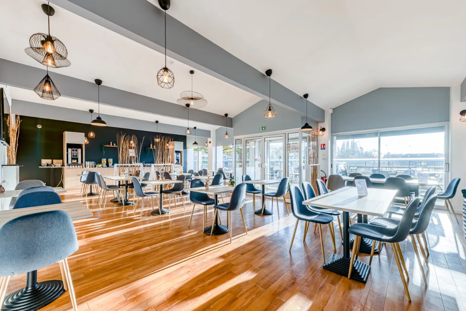 Bright, spacious restaurant-bar at Sure Hotel by Best Western Bordeaux Lac featuring wooden flooring, modern tables, blue chairs, large windows overlooking the lake, and gray ceiling beams with contemporary pendant lights.