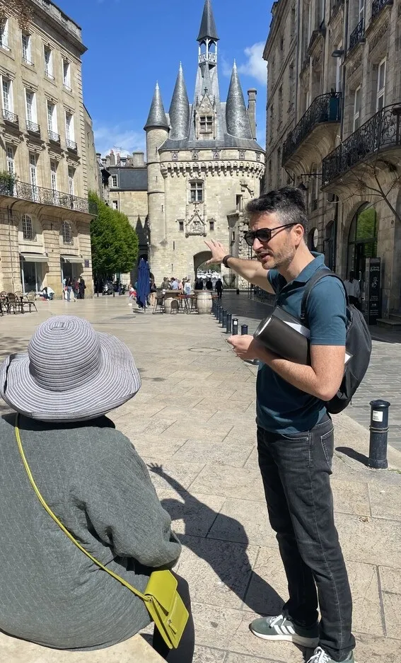 A tour guide speaking to a tourist wearing a white hat in front of the Porte Cailhau, a historic monument in Bordeaux.