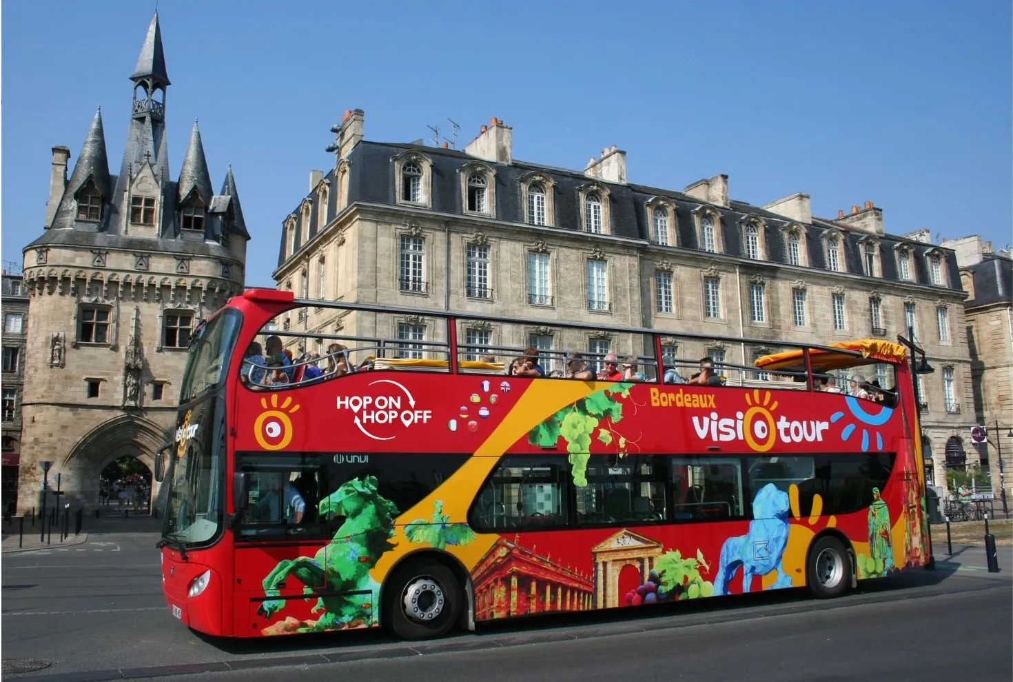 A red double-decker sightseeing bus from Bordeaux Visiotour (Hop On Hop Off) service parked in front of a medieval tower and Haussmann-style buildings in Bordeaux