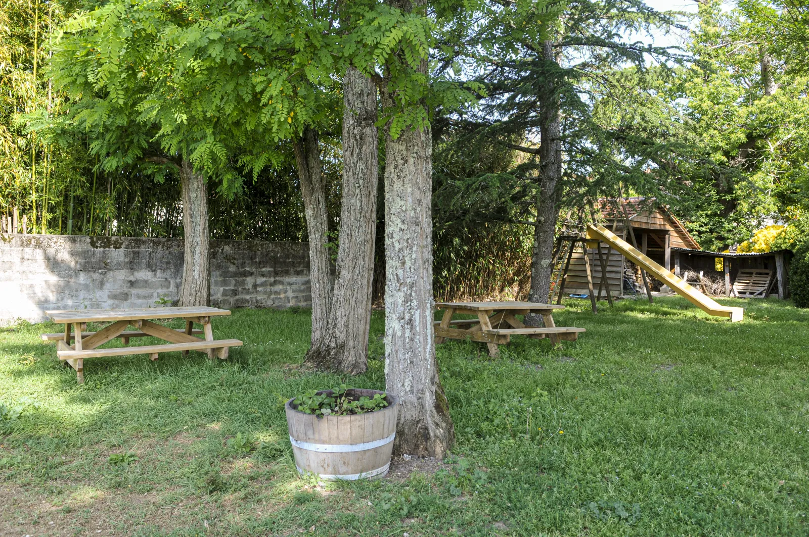Osmond Castle park in Bordeaux featuring wooden picnic tables, tall trees, a flower-filled barrel planter, stone wall, and playground structure