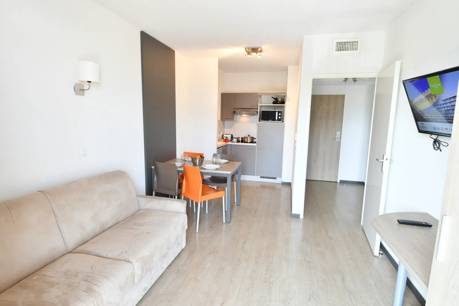 Modern studio interior featuring a living area with beige sofa, equipped kitchen, dining table with orange chairs, wall-mounted TV, light wood flooring, and minimalist décor in Bordeaux