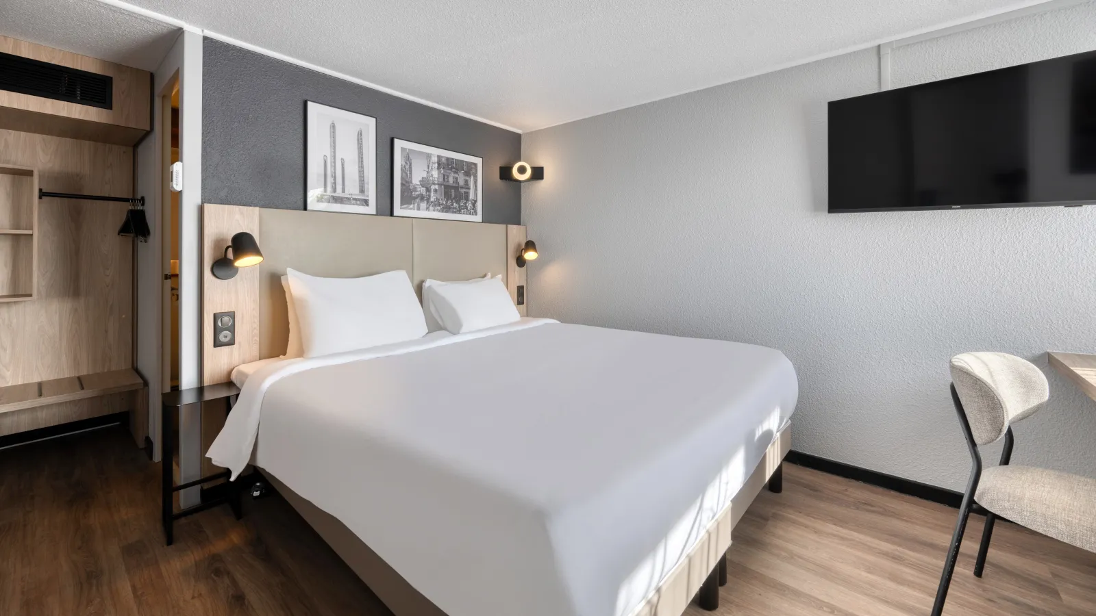Modern hotel room featuring a double bed with beige headboard, black and gold wall lights, wall-mounted TV, minimalist grey and white décor, and a light upholstered chair