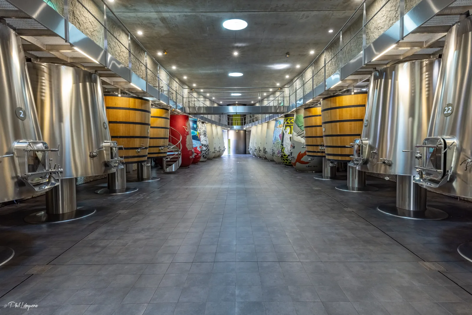 Interior of the winemaking cellar at Château les Carmes Haut-Brion in Bordeaux featuring stainless steel tanks and oak barrels lined on both sides of a central corridor
