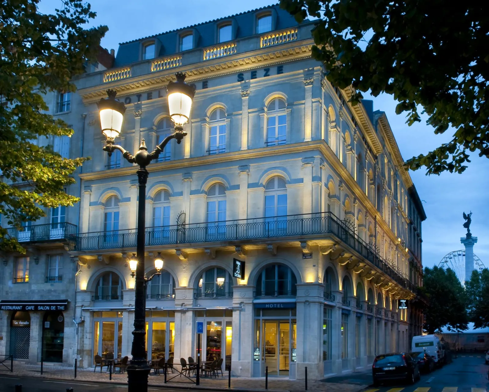 Illuminated facade of the Hôtel de Sèze in Bordeaux, a historic Haussmann-style building featuring columns, wrought-iron balconies and mansard roof, photographed at dusk with a street lamp in the foreground.