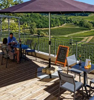 Wooden terrace of a wine estate with shade umbrella overlooking Bordeaux vineyards, guests seated, menu board displayed