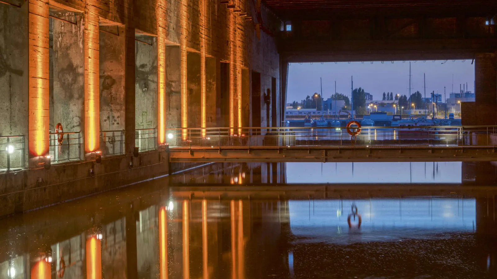 Night view of Bordeaux's submarine base featuring illuminated orange columns reflected in still water, overlooking the port and Garonne river at dusk.