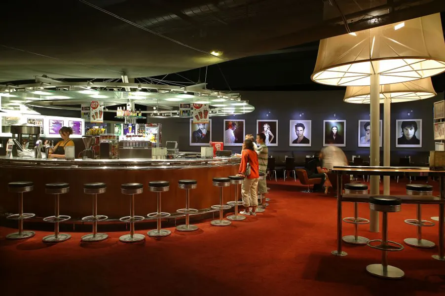 Interior of UGC Ciné Cité cinema in Bordeaux featuring a concession counter, bar stools, red carpet, illuminated portrait displays on walls, and waiting customers.