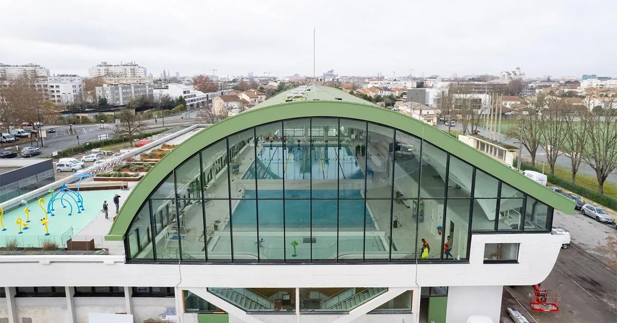 Aerial view of Galin Swimming Pool in Bordeaux, featuring a modern arched green roof, indoor pools visible through large glass windows, an outdoor blue pool on the left, surrounded by residential buildings and green spaces.
