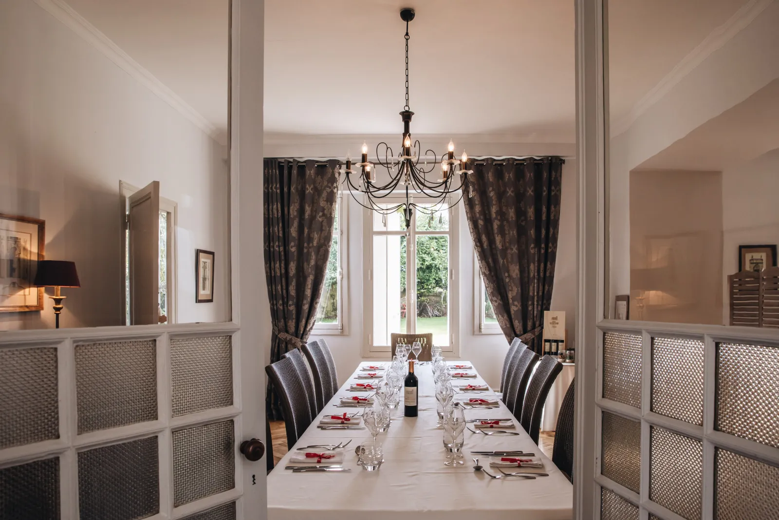 Elegant dining room at Château Ambe Tour Pourret in Bordeaux featuring a long formal table, wrought iron chandelier, brown curtains and glass doors with geometric panels