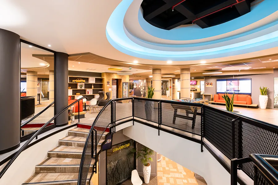 Modern lobby of Ibis Bordeaux Centre Mériadeck hotel featuring central staircase, black railings, luminescent blue circular ceiling, gray columns, orange furniture and reception areas
