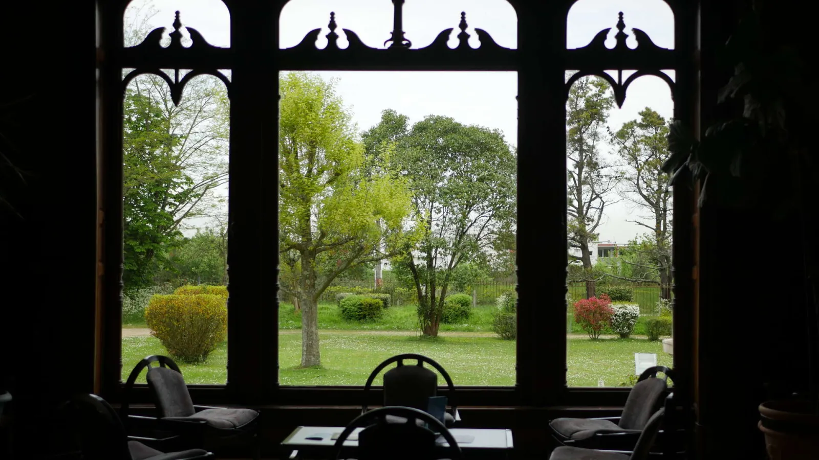 Interior view through three large black-framed windows with decorative tops overlooking a lush green garden with trees and flowering bushes, with black chairs in the foreground